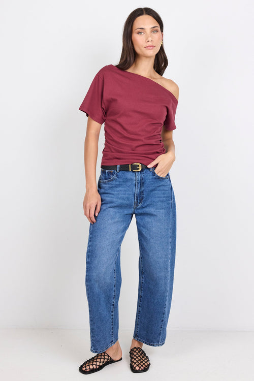 A woman modeling a burgundy one-shoulder short-sleeve t-shirt paired with blue jeans, a black belt, and black open-toe flat shoes, standing against a plain white studio background.