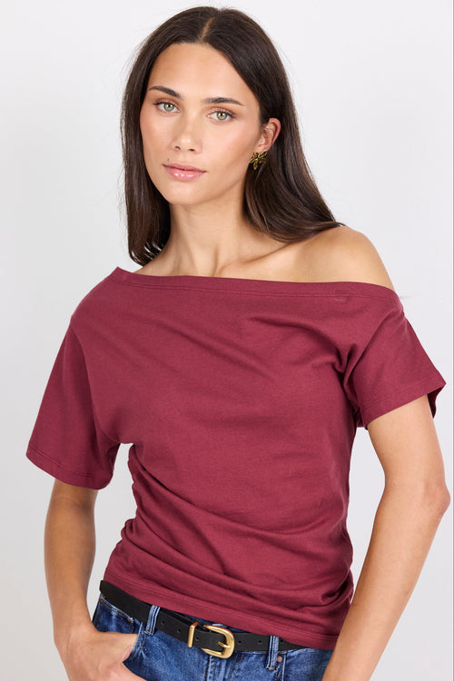 A woman modeling a short-sleeve off-the-shoulder t-shirt in burgundy, paired with blue jeans and a black belt, against a plain light gray background.