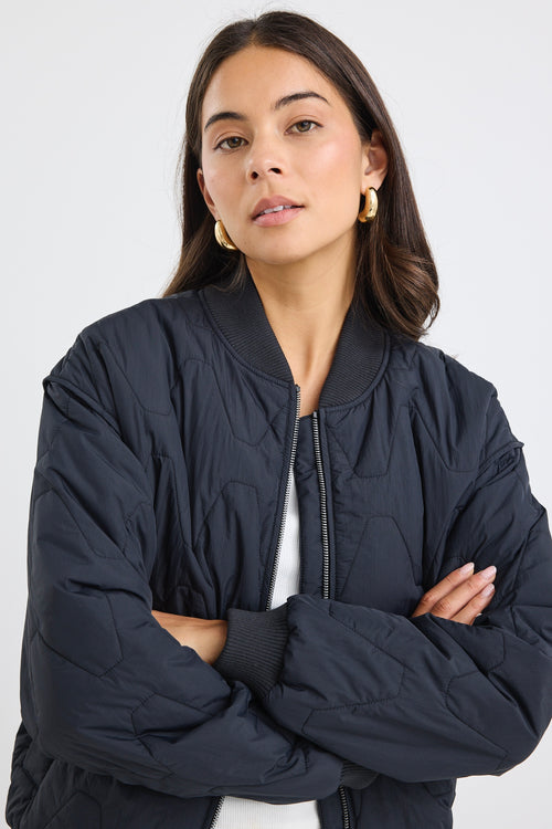 Woman wearing a black quilted zip-up puffer jacket with relaxed fit and ribbed trim, paired with light blue jeans and gold hoop earrings.