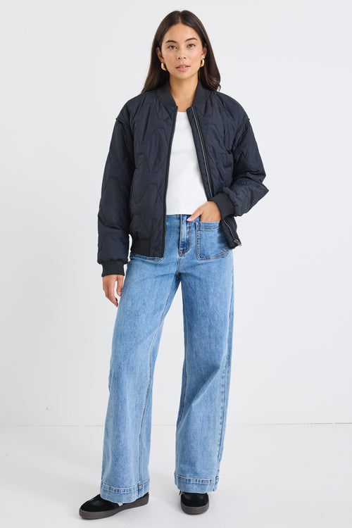 Woman wearing a black quilted zip-up puffer jacket with relaxed fit and ribbed trim, paired with light blue jeans and gold hoop earrings.