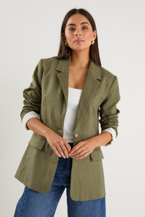 Woman wearing an olive green blazer over a white top and blue jeans on a plain background