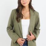 Woman wearing an olive green blazer over a white top and blue jeans on a white background