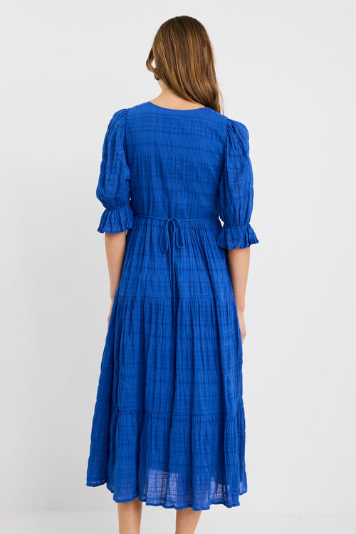 Woman wearing a bright azure blue tiered maxi dress with puff sleeves, button-front bodice, and v-neckline, styled with black woven flats.