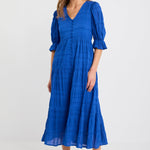 Woman wearing a bright azure blue tiered maxi dress with puff sleeves, button-front bodice, and v-neckline, styled with black woven flats.