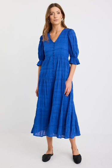 Woman wearing a bright azure blue tiered maxi dress with puff sleeves, button-front bodice, and v-neckline, styled with black woven flats.