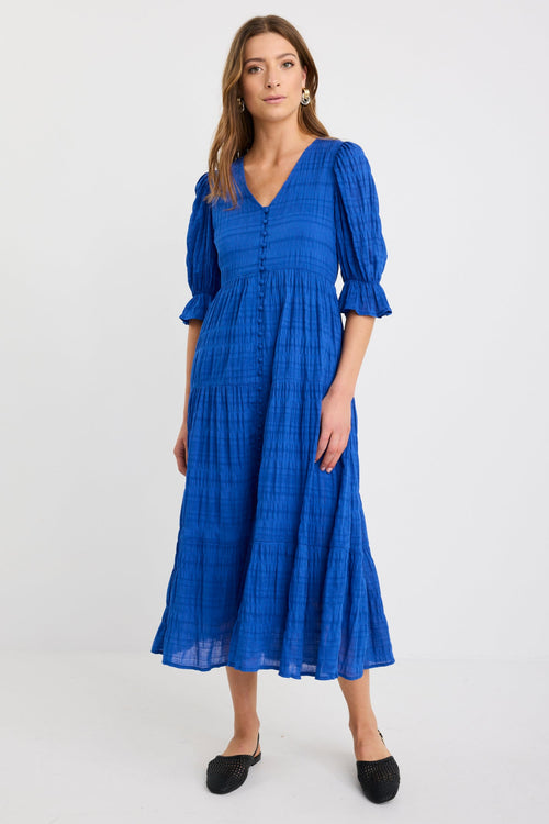Woman wearing a bright azure blue tiered maxi dress with puff sleeves, button-front bodice, and v-neckline, styled with black woven flats.