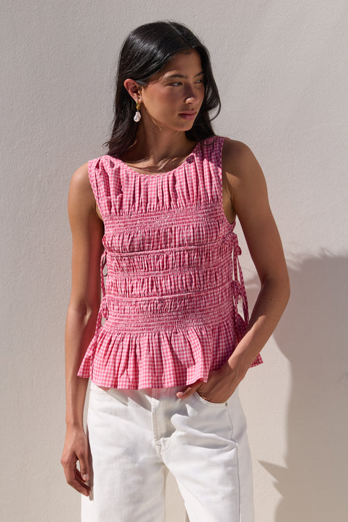 A woman wearing a pink and white gingham smocked sleeveless top with a ruffled hem, paired with white pants, standing against a textured off-white wall.