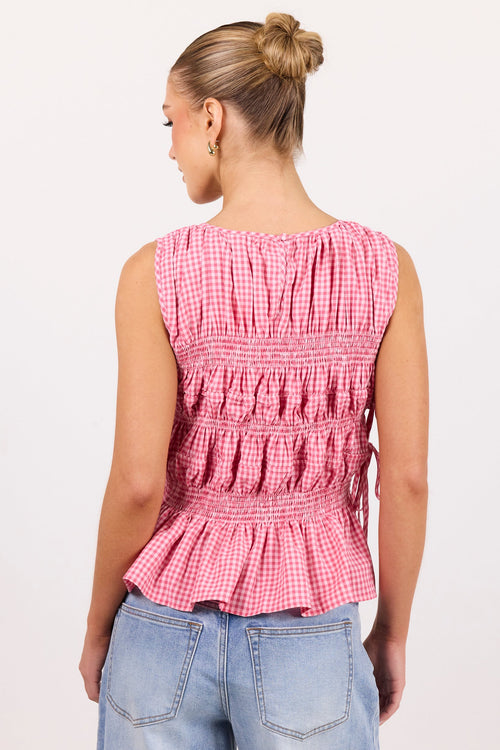 A woman shown from the back wearing a sleeveless pink and white gingham top with horizontal smocking and a ruffled peplum hem, paired with light blue jeans, against a plain white studio background.