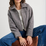 Woman wearing a gray jacket and blue jeans sitting on a brown stool against a white background