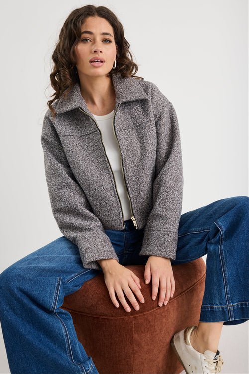 Woman wearing a gray jacket and blue jeans sitting on a brown stool against a white background