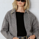 Woman wearing a gray jacket, black top, and blue jeans against a white wall.