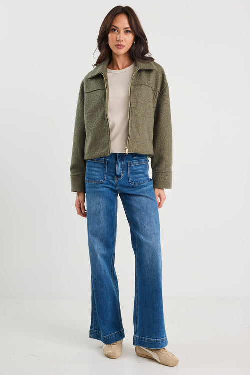 Woman wearing a green jacket, beige top, and blue jeans on a white background