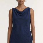 A model wearing a sleeveless navy blue cowl-neck top and matching navy pleated trousers, photographed against a plain off-white studio background.