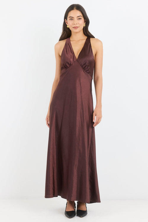 A woman modeling a sleeveless, floor-length halter gown in a deep plum-brown satin fabric, standing against a plain white studio background.