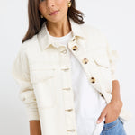 A woman modeling a cream-colored denim jacket with gold-tone buttons over a white t-shirt and blue jeans, set against a plain white studio background.