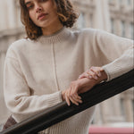 Person wearing a beige sweater standing in front of a building with a blurred red bus in the background