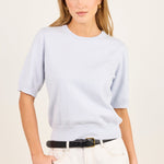 A woman modeling a light blue short-sleeve crewneck knit top, paired with cream-colored pants and a black belt, against a plain off-white studio background.