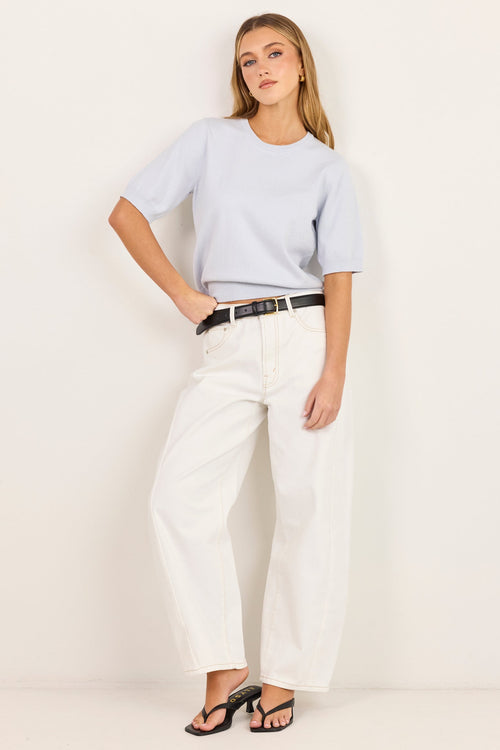 A woman modeling a light blue short-sleeve t-shirt, white wide-leg trousers, a black belt, and black sandals against a plain off-white studio background.