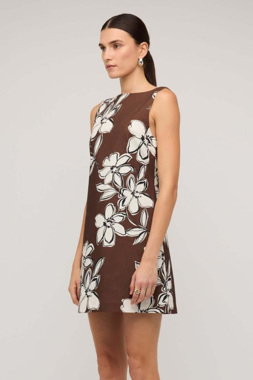Brown sleeveless mini dress with a straight neckline and bold white floral print, styled with a loose A-line fit, worn by a model holding a woven handbag.
