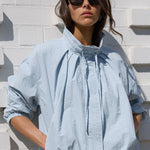 A woman wearing a light blue oversized crinkled blouse and white trousers, standing against a white textured brick wall.