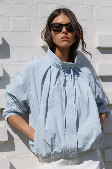 A woman wearing a light blue oversized crinkled blouse and white trousers, standing against a white textured brick wall.