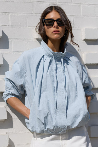 A woman wearing a light blue oversized crinkled blouse with a high gathered collar and white pants, standing against a white textured brick wall.