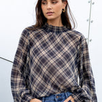 Woman wearing a plaid shirt and blue jeans standing against a white wall.