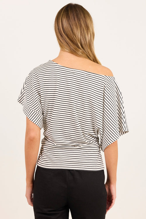 A woman viewed from the back wearing a black and white horizontally striped off-the-shoulder top and black pants, set against a plain off-white studio background.