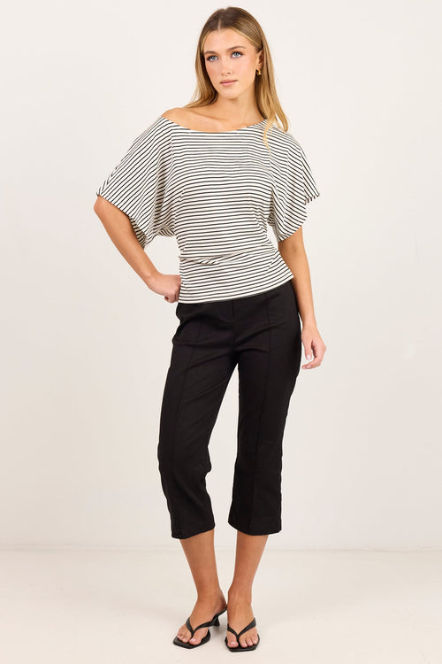 A woman modeling a black and white striped off-the-shoulder top with short flutter sleeves, paired with black cropped pants and black sandals, against a plain white studio background.