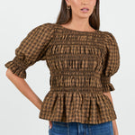 A woman wearing a chocolate and caramel checkered smocked top with puffed short sleeves, paired with high-waisted blue jeans.