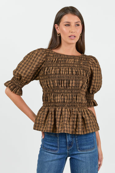 A woman wearing a chocolate and caramel checkered smocked top with puffed short sleeves, paired with high-waisted blue jeans.