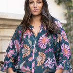 A woman wearing a dark teal floral blouse with pink, purple, and orange flowers, paired with blue jeans and a black belt, standing outdoors in front of a stone wall and greenery.