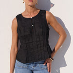 A woman modeling a sleeveless black textured top with vertical pleats and a button placket, paired with light blue jeans, standing against a white wall with a strong shadow.