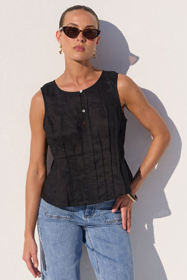 A woman modeling a sleeveless black textured top with vertical pleats and a button placket, paired with light blue jeans, standing against a white wall with a strong shadow.