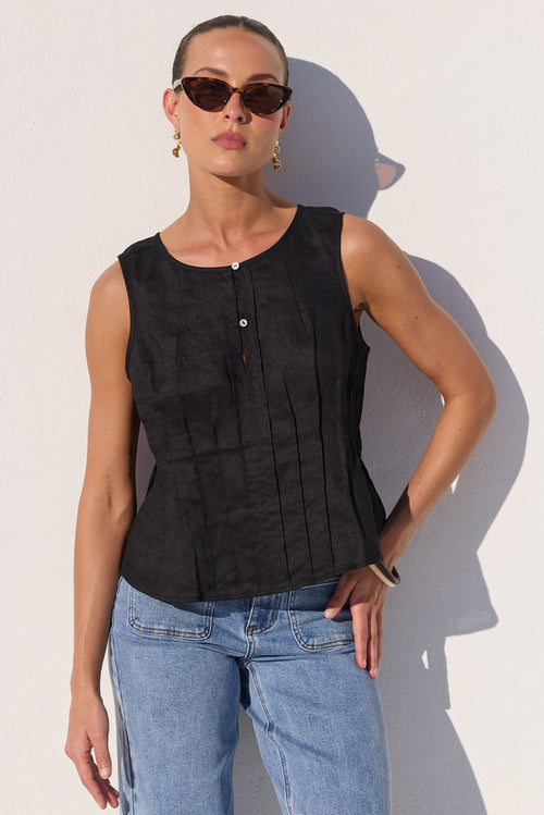 A woman modeling a sleeveless black textured top with vertical pleats and a button placket, paired with light blue jeans, standing against a white wall with a strong shadow.
