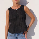 A woman wearing a black sleeveless top with vertical pleats and light blue jeans, standing against a white wall with a strong shadow.