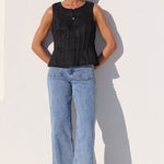 A woman stands against a white wall wearing a sleeveless black top, light blue wide-leg jeans, and black sandals.