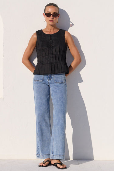 A woman stands against a white wall wearing a sleeveless black top, light blue wide-leg jeans, and black sandals.