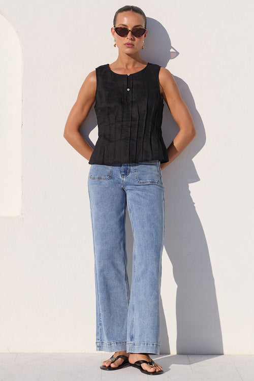 A woman stands against a white wall wearing a sleeveless black top, light blue wide-leg jeans, and black sandals.
