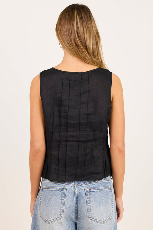 A woman viewed from the back wearing a sleeveless black top with vertical pleats and light-wash blue jeans, against a plain off-white studio background.