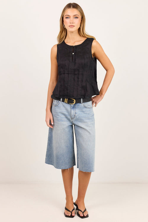 A woman modeling a sleeveless black textured top paired with light-wash denim culottes and black sandals, standing against a plain off-white studio background.