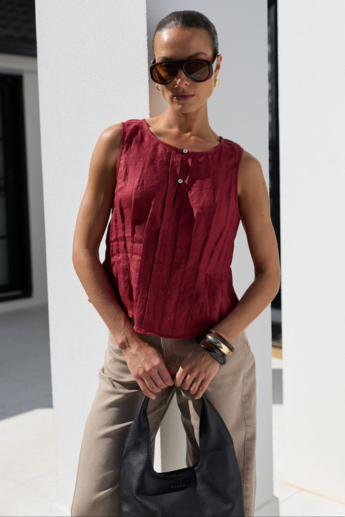 A woman wearing a sleeveless deep red top and beige trousers, holding a black leather bag, standing in front of a white architectural background.