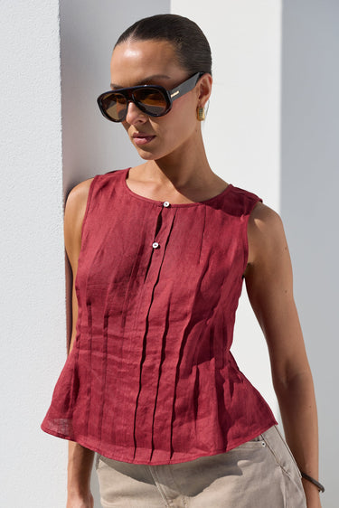 A woman wearing a sleeveless, deep red linen peplum top with vertical pleats and a button detail, paired with beige trousers and sunglasses, standing against a white wall.