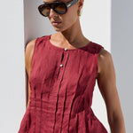 A woman wearing a sleeveless, peplum-style top in a deep burgundy red, paired with beige trousers and dark sunglasses, standing against a white wall.