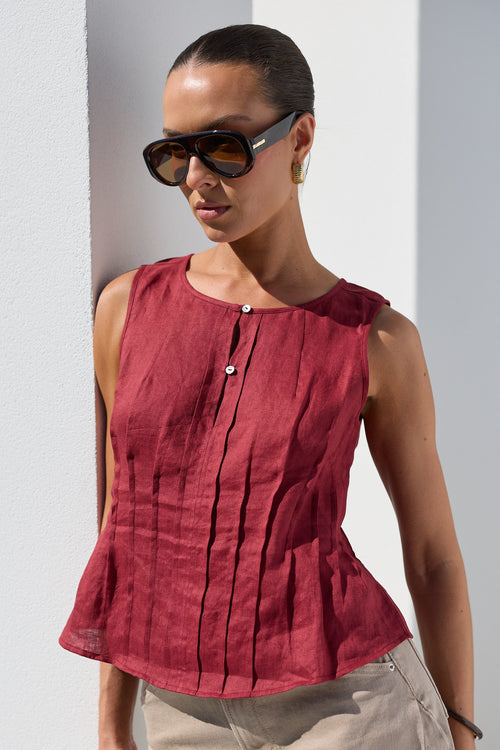 A woman wearing a sleeveless, peplum-style top in a deep burgundy red, paired with beige trousers and dark sunglasses, standing against a white wall.