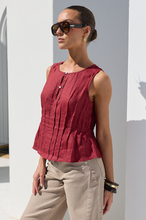 A woman wearing a sleeveless, deep red peplum top and beige trousers, standing in a bright white architectural setting with columns and casting a shadow on the wall.