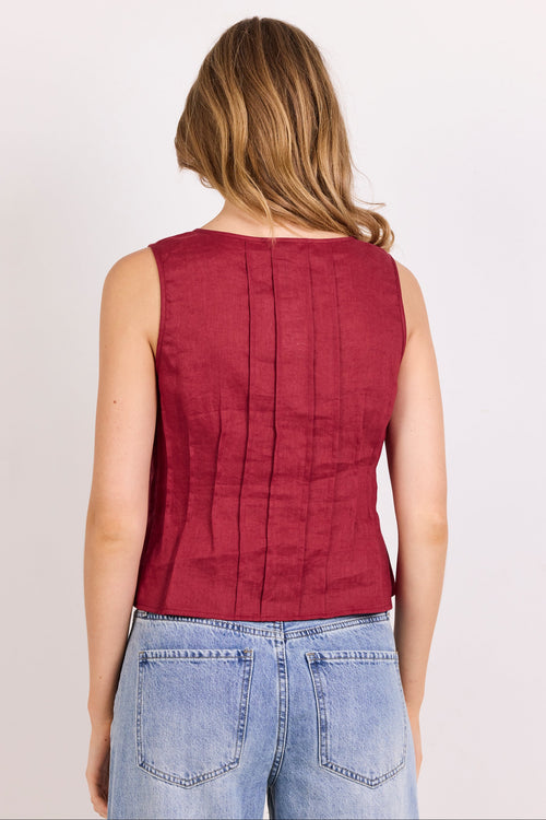A woman viewed from the back wearing a sleeveless burgundy top with vertical pleats and light blue jeans, set against a plain white background.