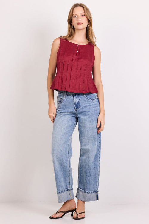 A woman modeling a sleeveless burgundy top with vertical pleats and a button placket, paired with light-wash blue jeans and black sandals, against a plain white studio background.