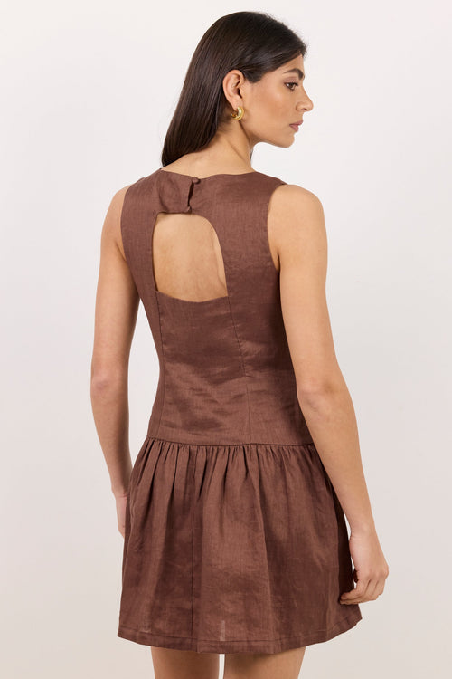 A woman modeling a sleeveless brown mini dress from the back, showing a keyhole cutout at the upper back and a gathered skirt, against a plain off-white studio background.