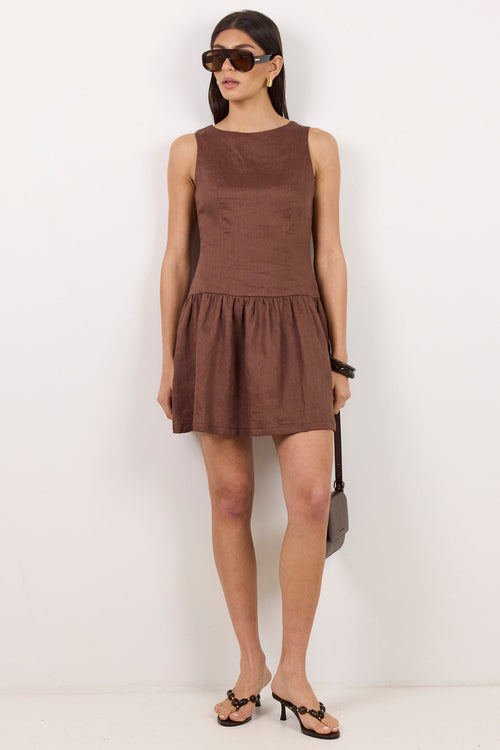 A woman modeling a sleeveless brown mini dress with a gathered waist, paired with black sandals, sunglasses, and a small dark handbag, standing against a plain white wall.